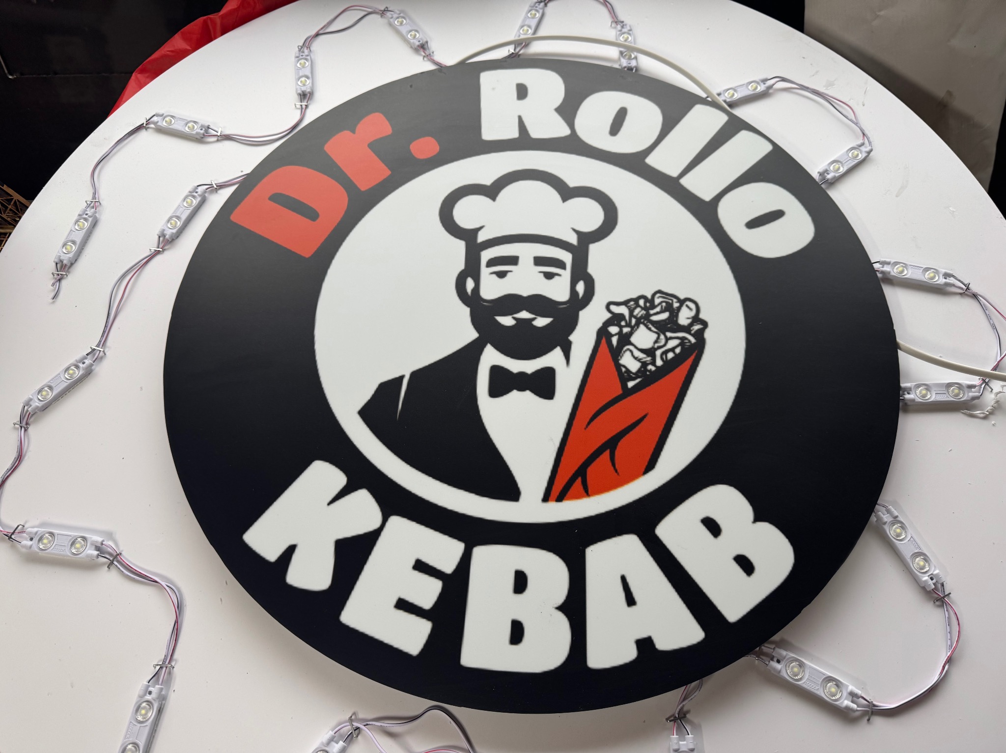 Okrągły, podświetlany szyld reklamowy 'Dr. Rollo Kebab' z wizerunkiem kucharza i daniem kebab, otoczony diodami LED na białym tle.