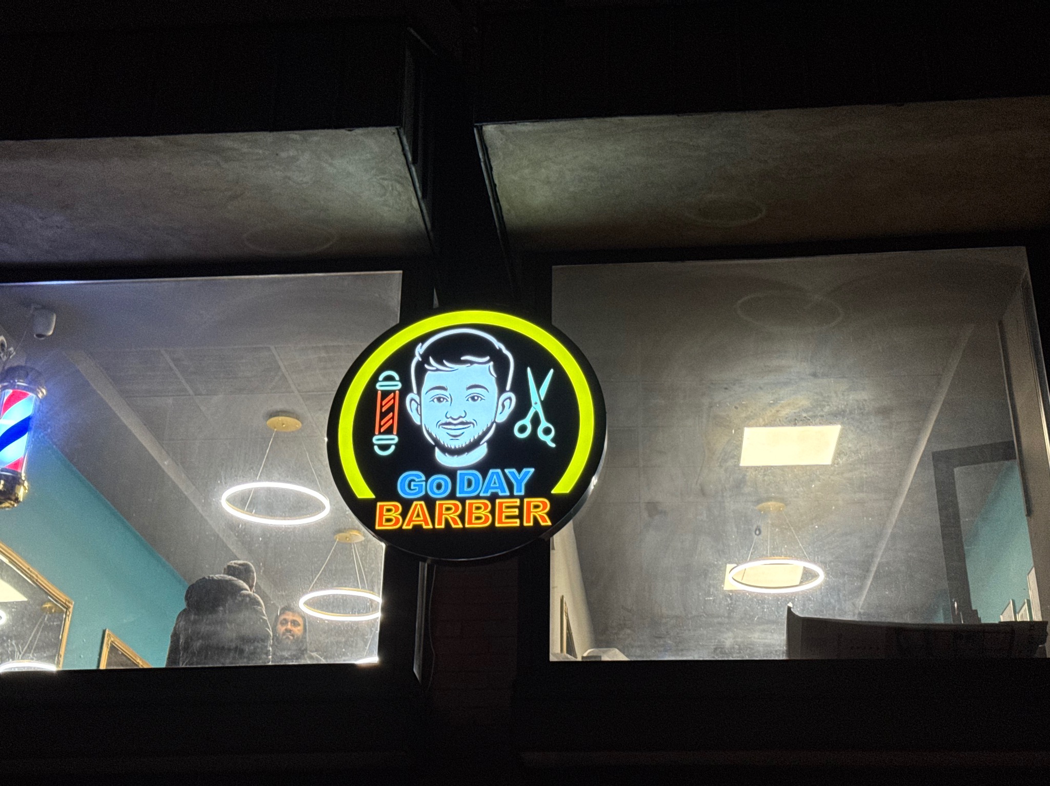Neonowy szyld 'Go Day Barber' z wizerunkiem mężczyzny i nożyczkami, widoczny przez okno salonu z klientami i nowoczesnym oświetleniem.