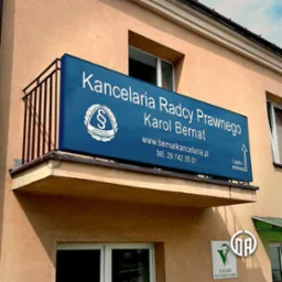 Niebieski szyld z białym napisem 'Kancelaria Radcy Prawnego Karol Bernat' umieszczony na balkonie budynku, widoczny znak Krajowej Izby Radców Prawnych oraz adres strony internetowej i numer telefonu.