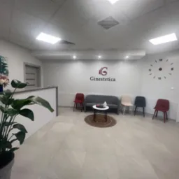 Jasne, nowoczesne wnętrze poczekalni z rośliną doniczkową, obrazem kobiety z kwiatami, minimalistycznym logo firmy 'Ginestetica' na ścianie i zegarem ściennym.