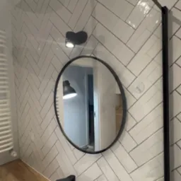 Nowoczesna łazienka z białą umywalką, czarną armaturą i lustrem w okrągłej ramie na ścianie wyłożonej białymi płytkami w jodełkę, oświetlona punktowym reflektorem.