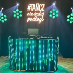 Oświetlona konsola DJ-a z neonowym napisem '#TANCZ nie żałuj podłogi', głośniki RCF i reflektory tworzą klubową atmosferę. Laptop na konsoli.
