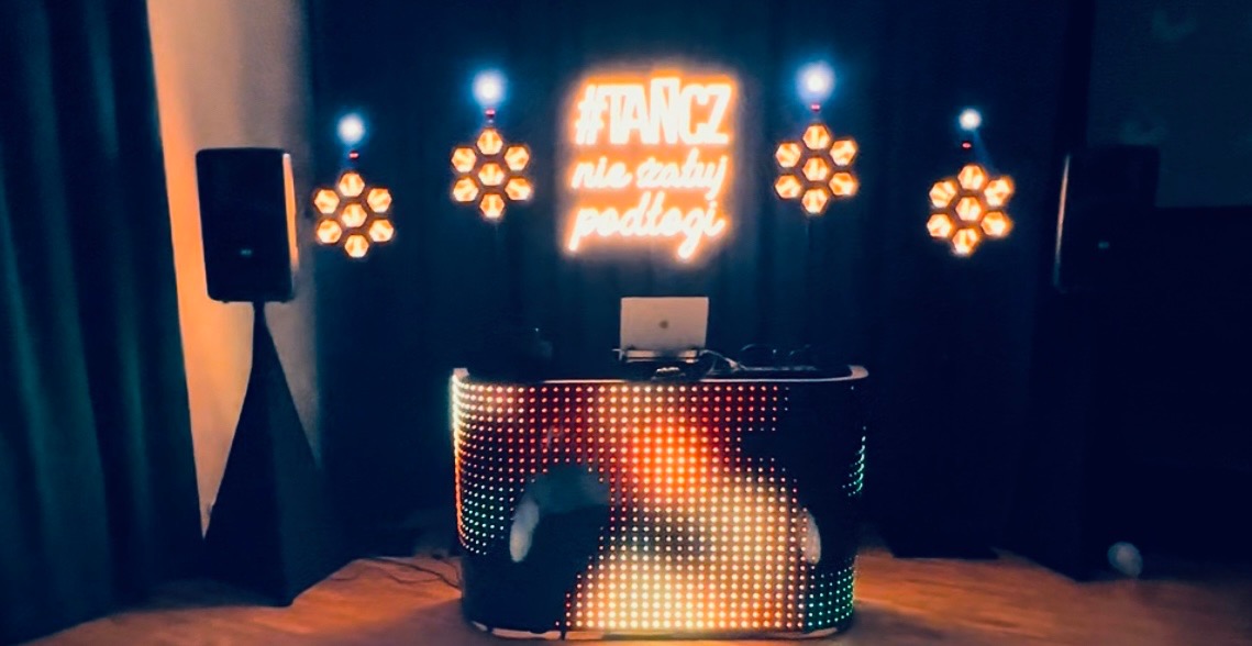 Podświetlony stół DJ-ski z laptopem, głośnikami i neonem '#TANCZ nie stały podłogi' tworzy klubową atmosferę. Dekoracyjne lampy w kształcie plastrów miodu.
