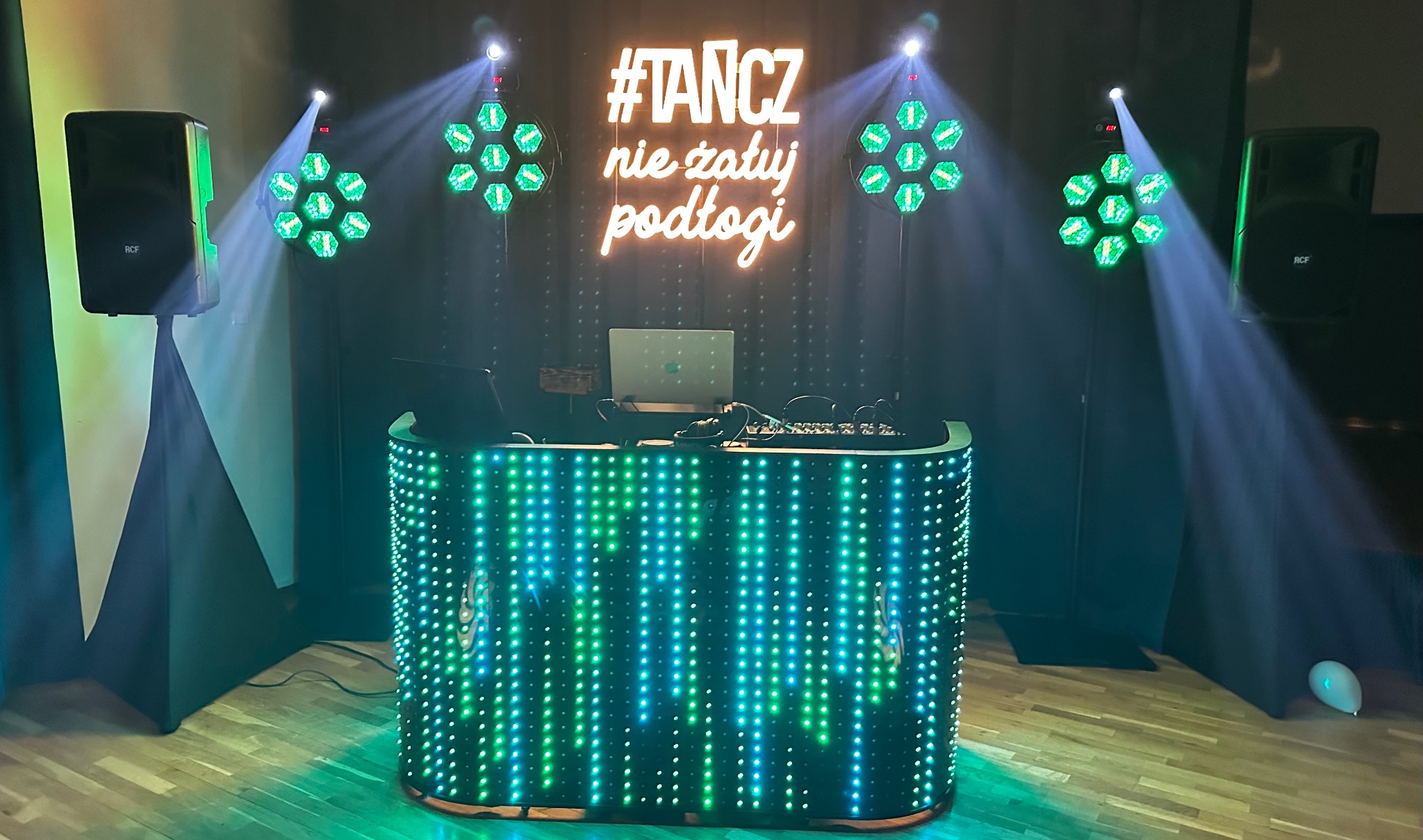 Oświetlona konsola DJ-a z neonowym napisem '#TANCZ nie żałuj podłogi', głośniki RCF i reflektory tworzą klubową atmosferę. Laptop na konsoli.