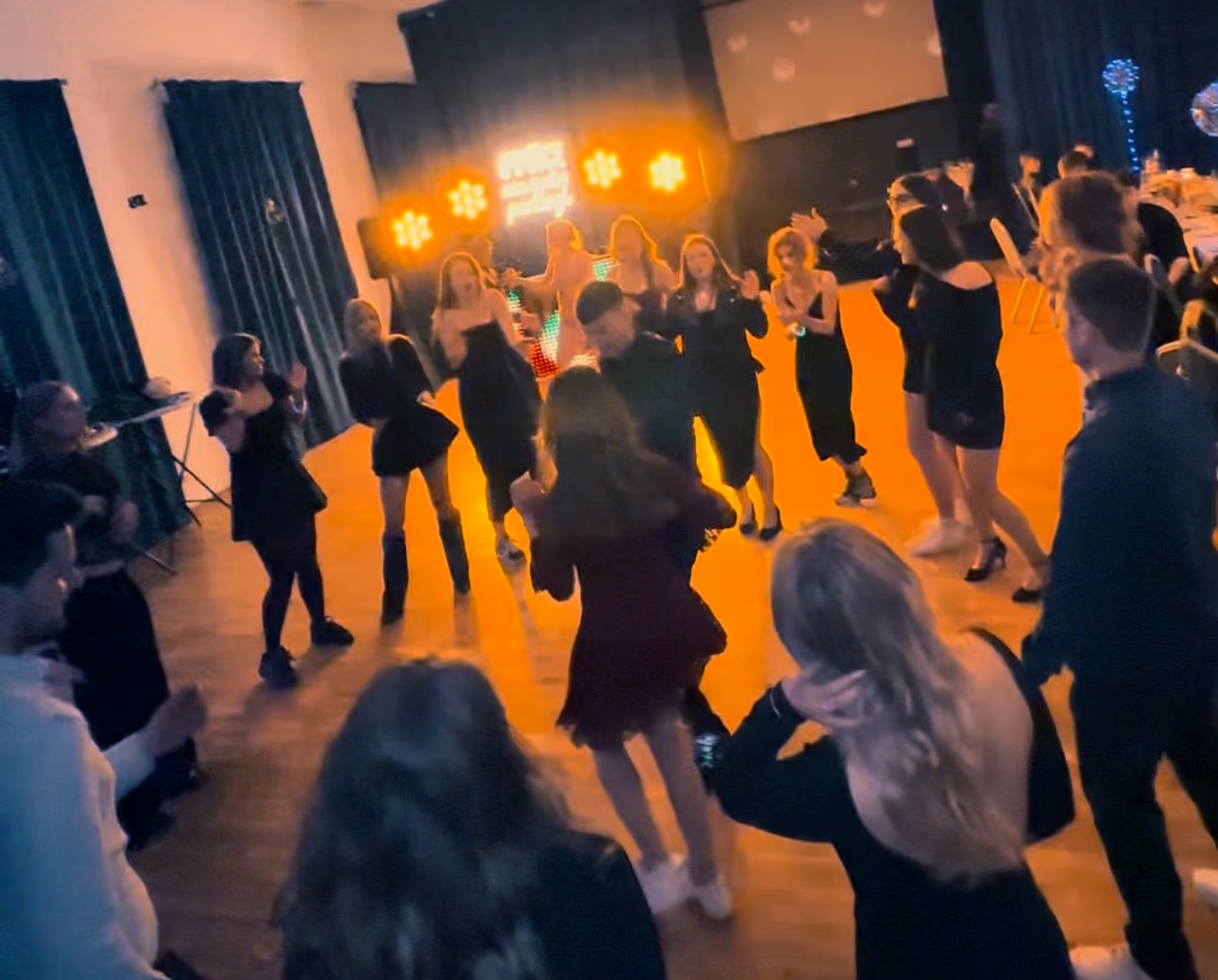 Zespół muzyczny na scenie podczas występu na imprezie. Wokół tańczą goście, oświetleni światłami reflektorów. Widoczny parkiet i elementy dekoracji.