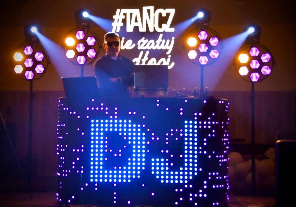 DJ w okularach za podświetlonym stanowiskiem z napisem 'DJ', oświetlonym reflektorami na tle napisu '#TANCZ nie żałuj dłoni'.