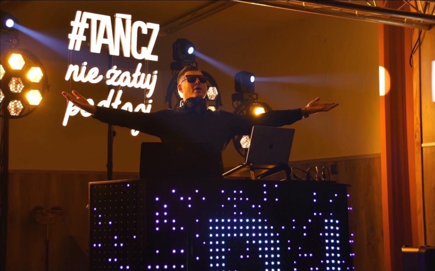 DJ w okularach za podświetlonym stanowiskiem, z rozłożonymi rękami, przed neonem z hasłem '#TANCZ nie żałuj podatków', oświetlony reflektorami.