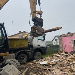 Usługi transportowePiotr Wężyk - Żuraw chwyta element z rozbieranego budynku, obok stoi ciężarówka. Widoczne gruzy i fragmenty konstrukcji. W tle domy i pochmurne niebo.