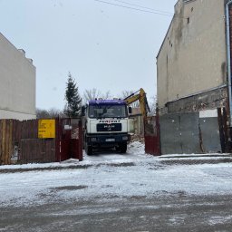 Usługi transportowePiotr Wężyk - Ciężarówka MAN na tle rozbiórki budynku w zimowej scenerii. Widoczny gruz i koparka. Tablica informacyjna budowy po lewej stronie. Śnieg na ulicy.