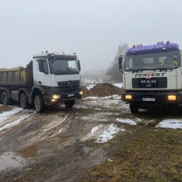 Usługi transportowePiotr Wężyk - Dwa samochody ciężarowe (Mercedes i MAN) na błotnistym terenie z resztkami śniegu, prawdopodobnie podczas prac ziemnych. Widoczny operator w kabinie MAN.