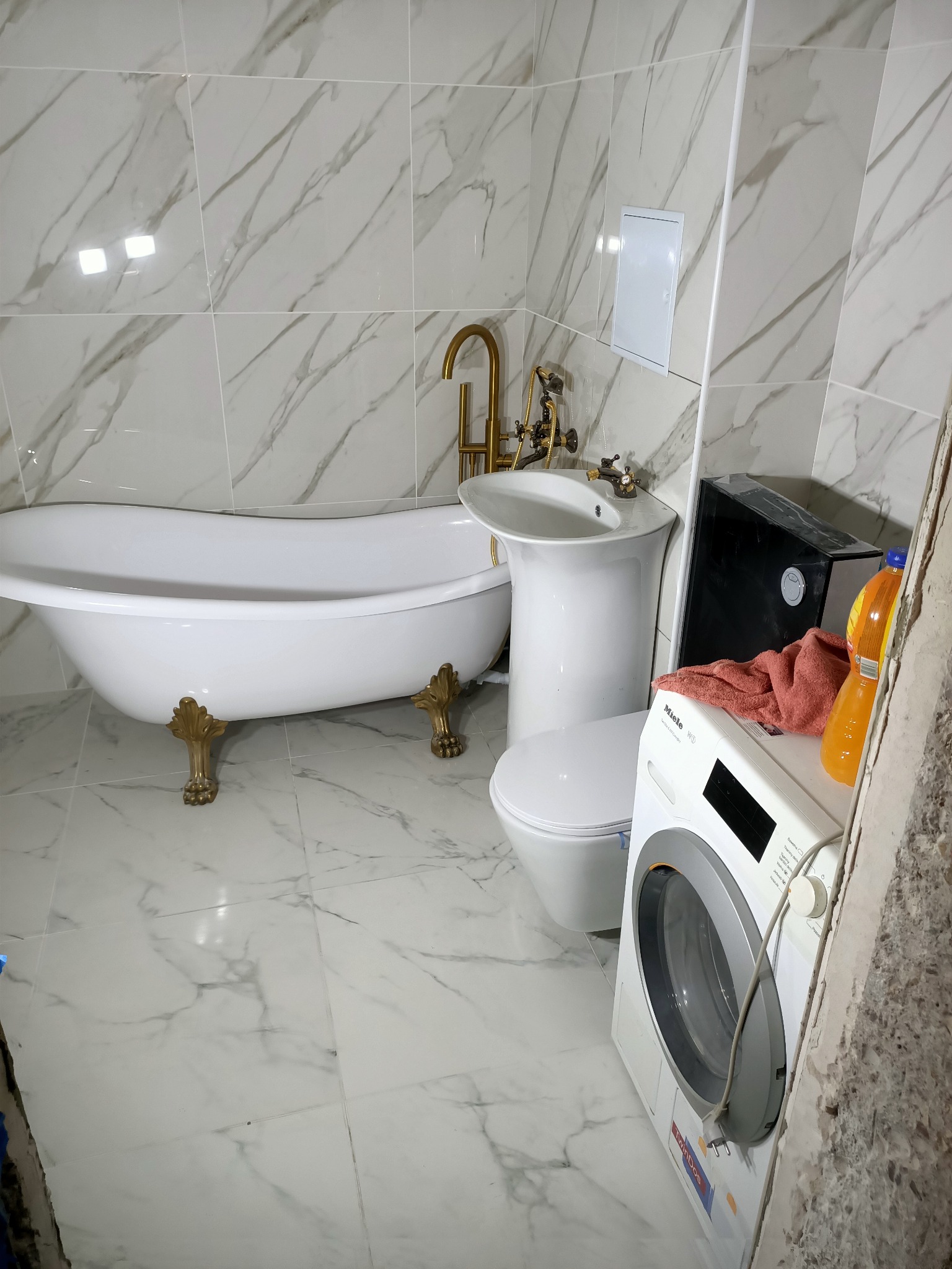 Biała wanna wolnostojąca na złotych nóżkach, umywalka na postumencie i toaleta w łazience wykończonej marmurowymi płytkami, widoczna pralka Miele i niedokończone wykończenie ściany.