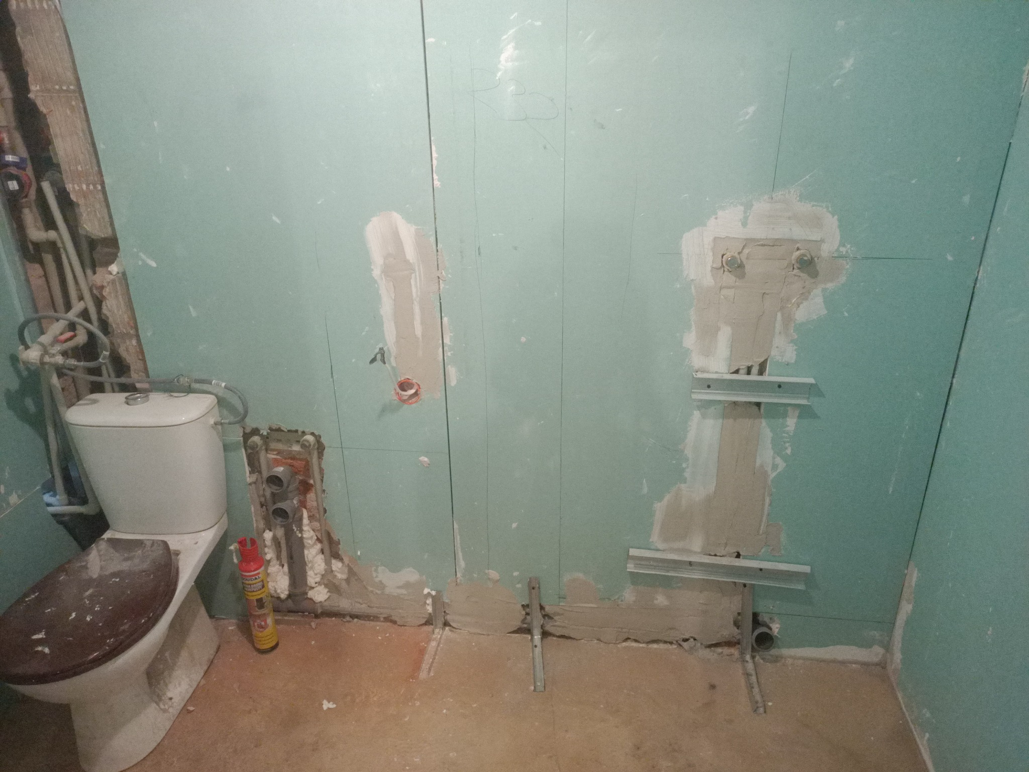 Łazienka w trakcie remontu: widoczne płyty gipsowo-kartonowe, instalacja wodno-kanalizacyjna, mocowania stelaża podtynkowego i częściowo zamontowana toaleta ze śladami zabrudzeń na desce sedesowej.