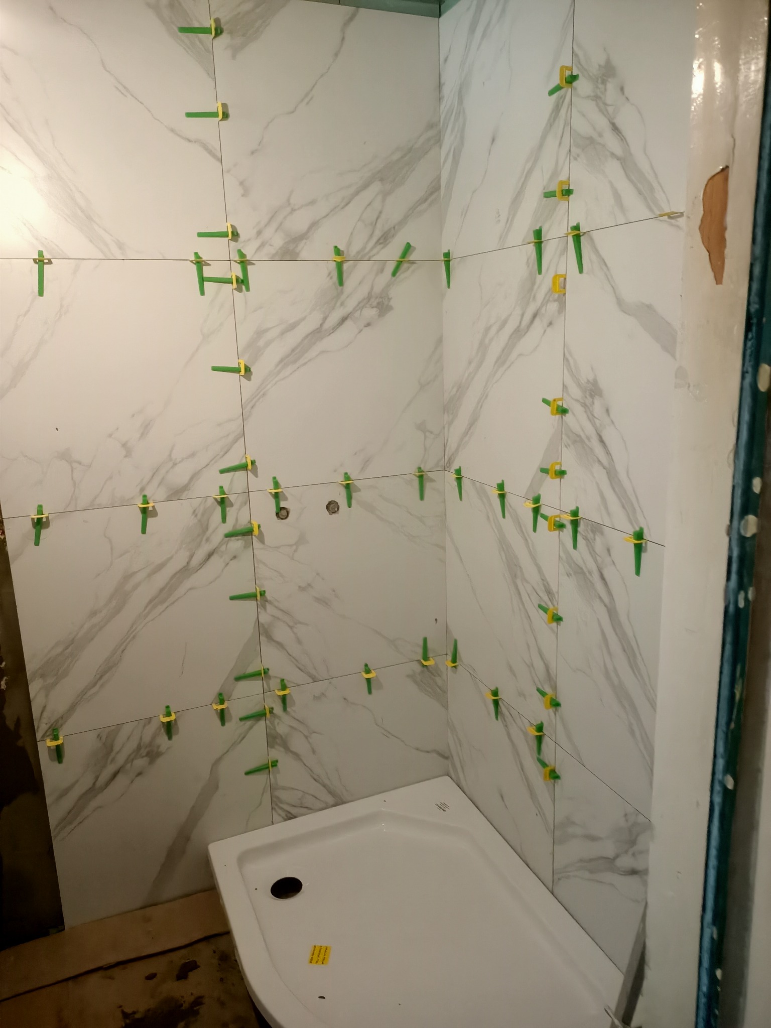 Fragment łazienki w trakcie wykańczania: białe płytki imitujące marmur z zielonymi krzyżykami dystansowymi i sznurkiem poziomującym, widoczny brodzik.
