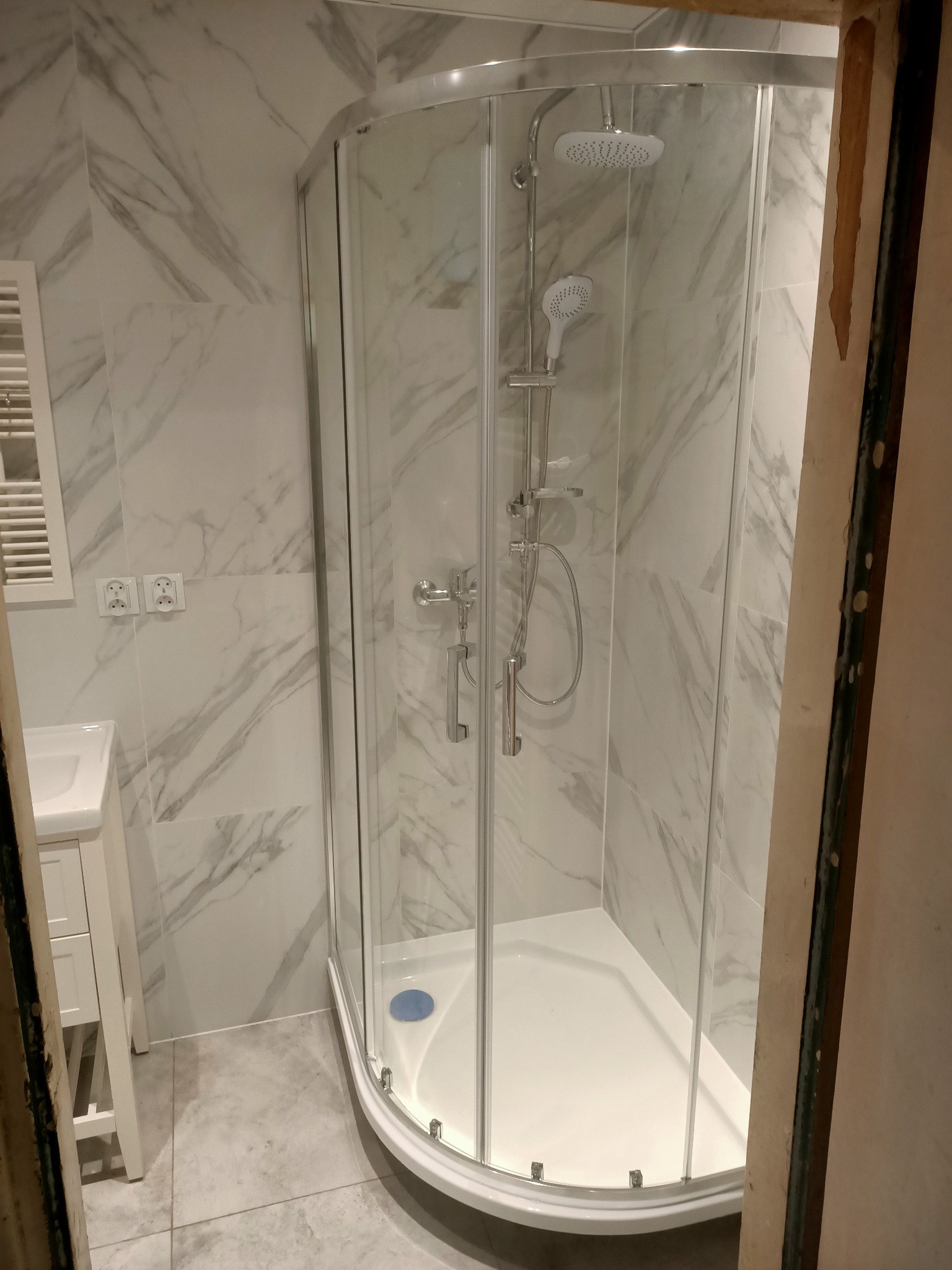 Nowoczesna kabina prysznicowa z hydromasażem, białe płytki imitujące marmur na ścianach, widoczne gniazdka elektryczne i grzejnik drabinkowy, fragment surowej ościeżnicy drzwiowej po prawej stronie.