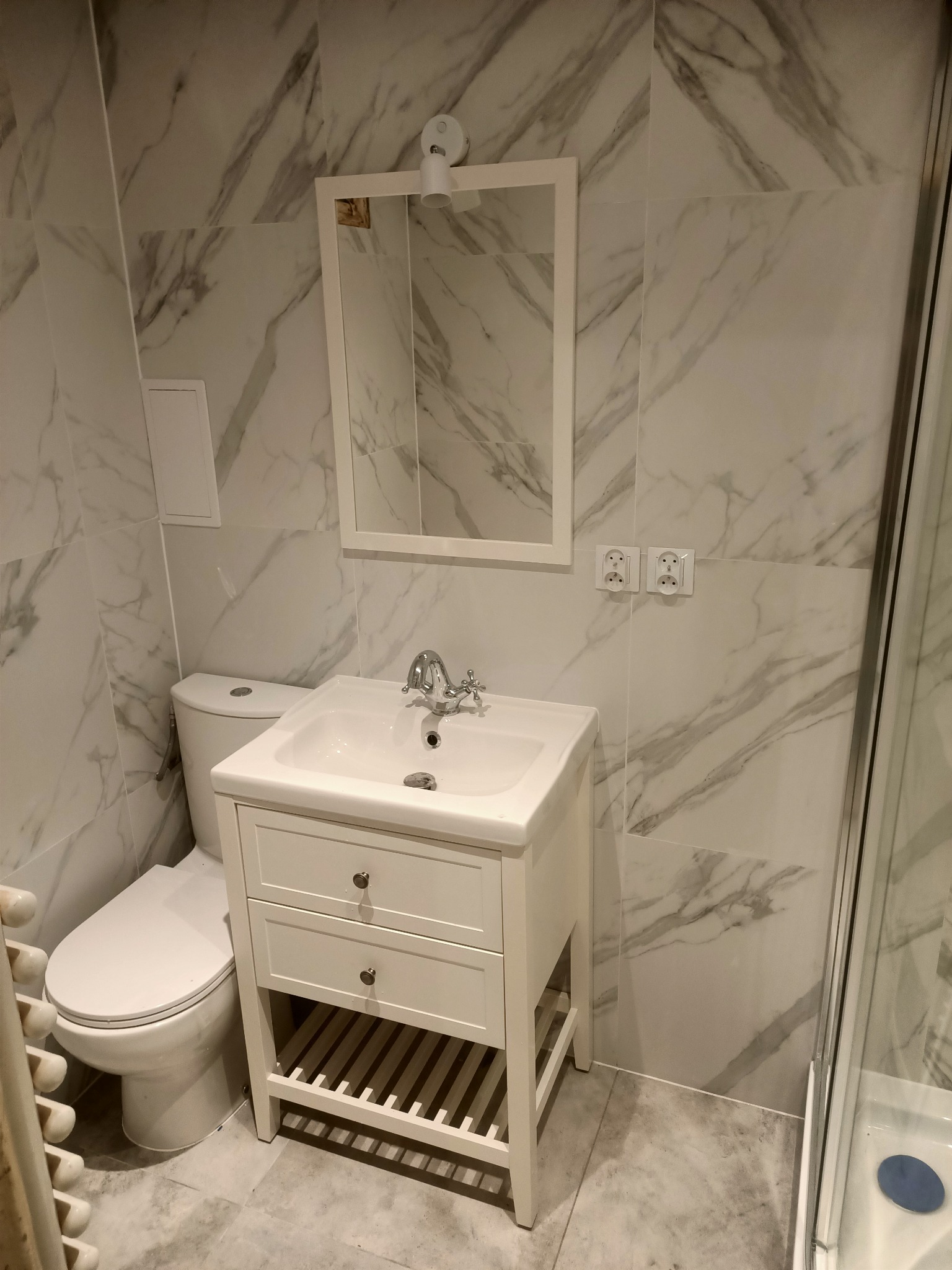 Biała umywalka z szafką i lustrem w łazience wykończonej marmurowymi płytkami, widoczne gniazdka elektryczne i fragment kabiny prysznicowej.