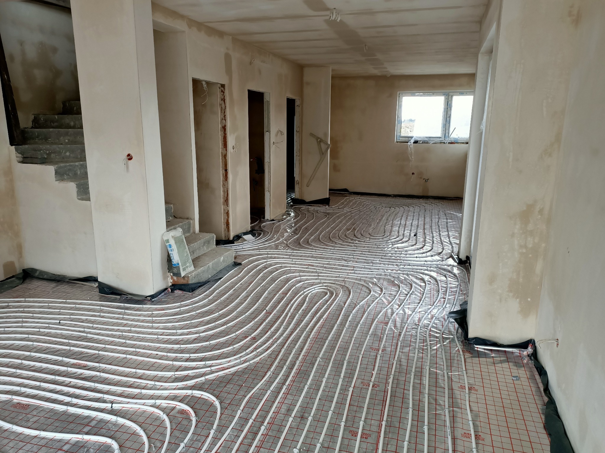 Instalacja ogrzewania podłogowego w surowym stanie, rury grzewcze ułożone na izolacji z czerwonymi liniami, ściany w trakcie prac wykończeniowych, widoczne schody i okno.