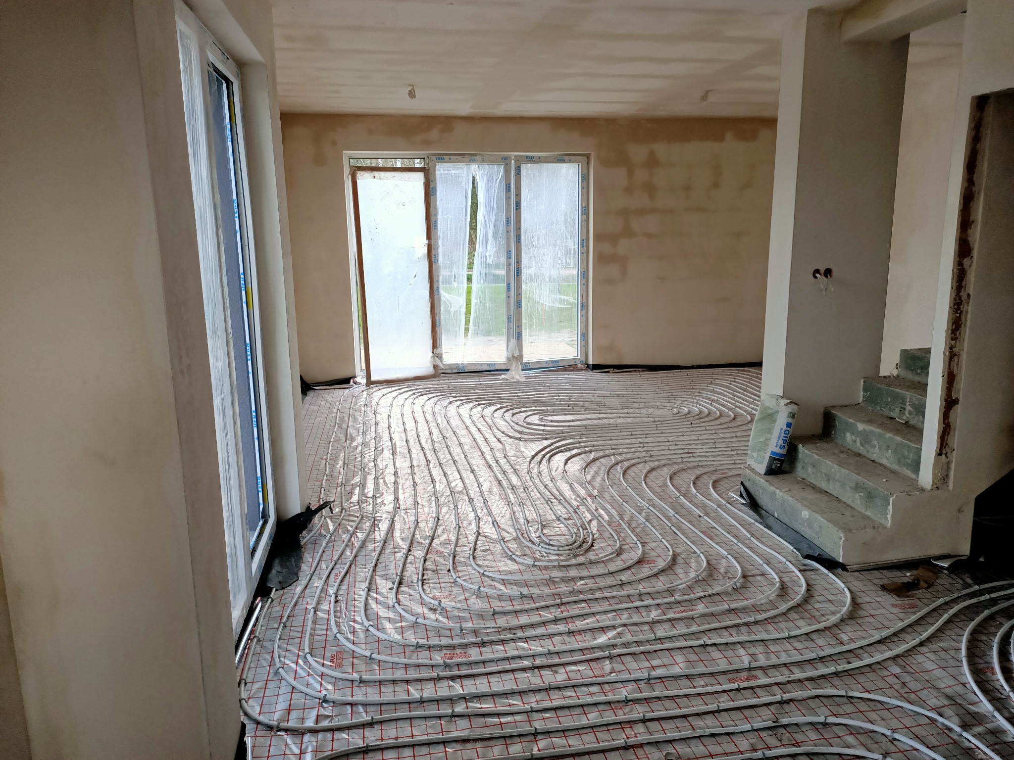 Rozłożone rury ogrzewania podłogowego na folii z siatką, widok na surowy stan ścian, okna i schody w trakcie wykończenia.