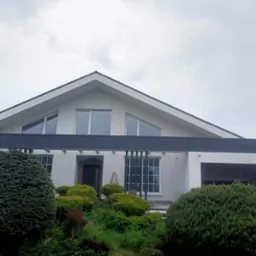 Elewacja nowoczesnego domu jednorodzinnego w Toruniu, z szarą belką nad wejściem i jasnymi ścianami, otoczona bujną zielenią.