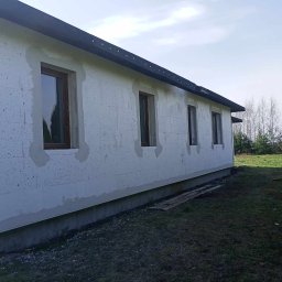 Rem-bud - Ściana budynku z szarego betonu komórkowego, z widocznymi brązowymi ramami okiennymi, częściowo przygotowana do dalszych prac wykończeniowych, widok z zewnątrz na tle trawnika i drzew.