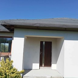 Rem-bud - Dom w trakcie budowy: surowa elewacja w kolorze białym, tymczasowe pokrycie dachu, brązowe okno z drewnianą ramą i nowoczesne drzwi wejściowe z metalowymi akcentami. Widoczna żółta roślina...