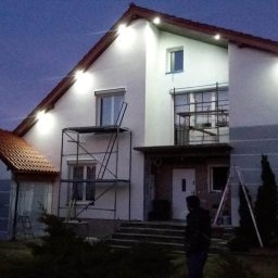 Rem-bud - Dwukondygnacyjny dom jednorodzinny w trakcie prac elewacyjnych, oświetlony reflektorami, z widocznymi rusztowaniami i fragmentem pomalowanej szarej elewacji z białymi pasami, sfotografowany...