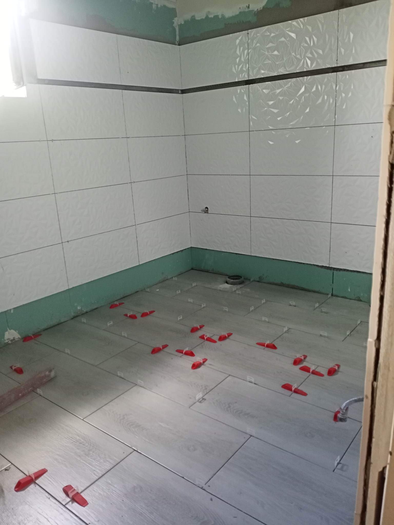 Łazienka w trakcie układania płytek podłogowych i ściennych w jasnych odcieniach szarości i bieli, z użyciem czerwonych klinów poziomujących, widoczne niedokończone wykończenia ścian.