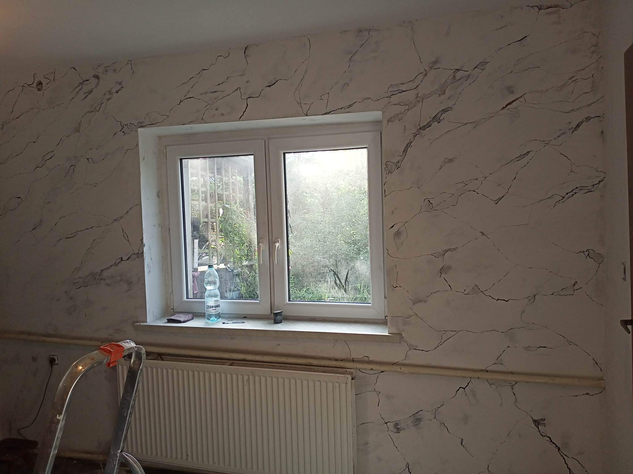 Ściana w pokoju z efektem marmuru, widoczne okno z widokiem na zieleń, grzejnik i fragment drabiny malarskiej.
