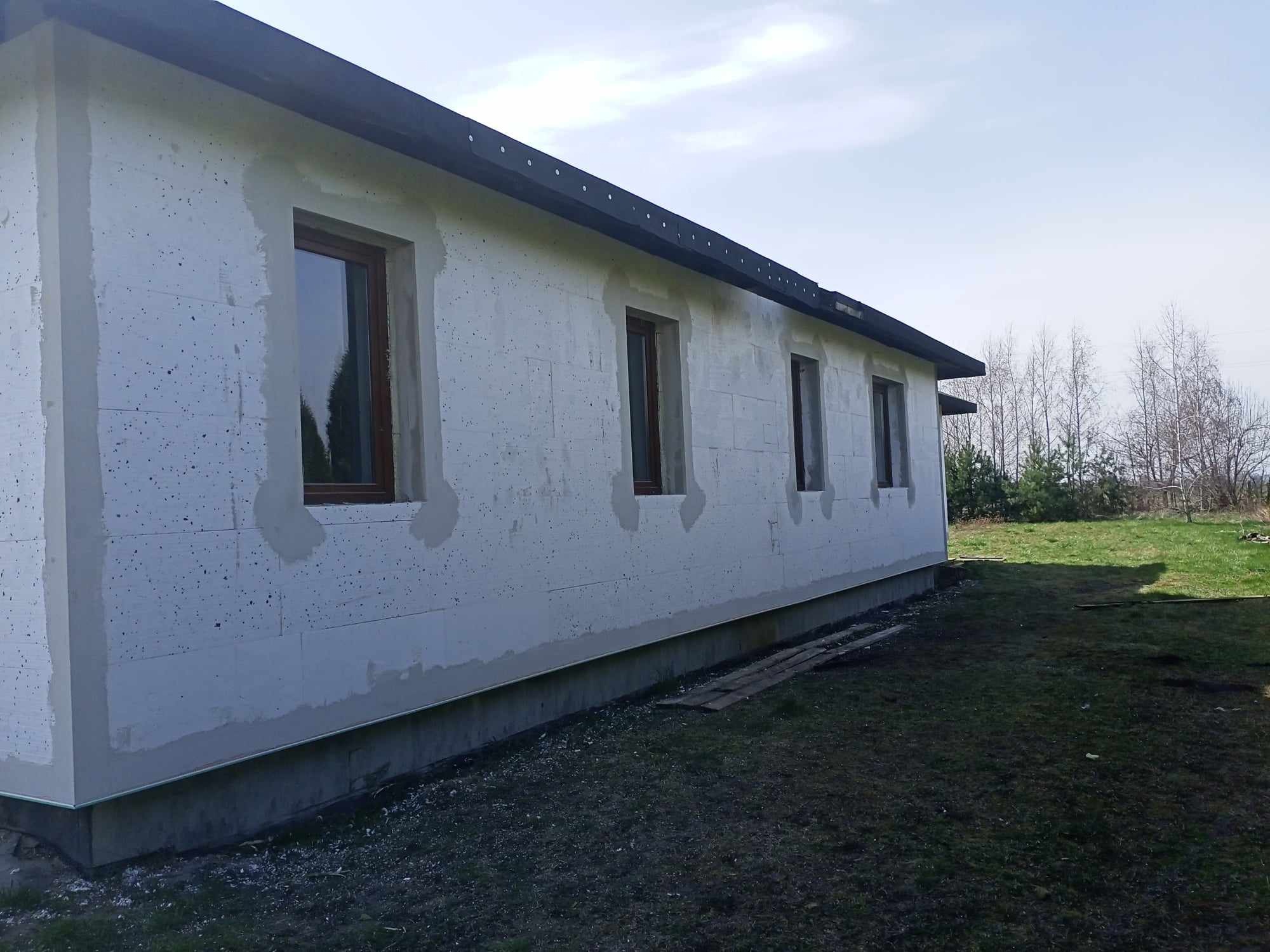 Ściana budynku z szarego betonu komórkowego, z widocznymi brązowymi ramami okiennymi, częściowo przygotowana do dalszych prac wykończeniowych, widok z zewnątrz na tle trawnika i drzew.