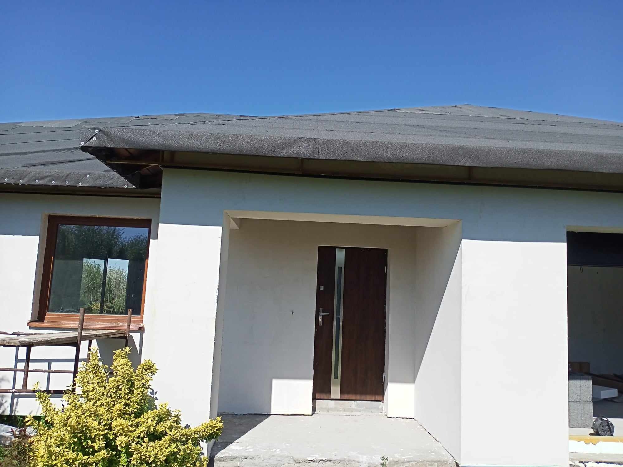 Dom w trakcie budowy: surowa elewacja w kolorze białym, tymczasowe pokrycie dachu, brązowe okno z drewnianą ramą i nowoczesne drzwi wejściowe z metalowymi akcentami. Widoczna żółta roślina...