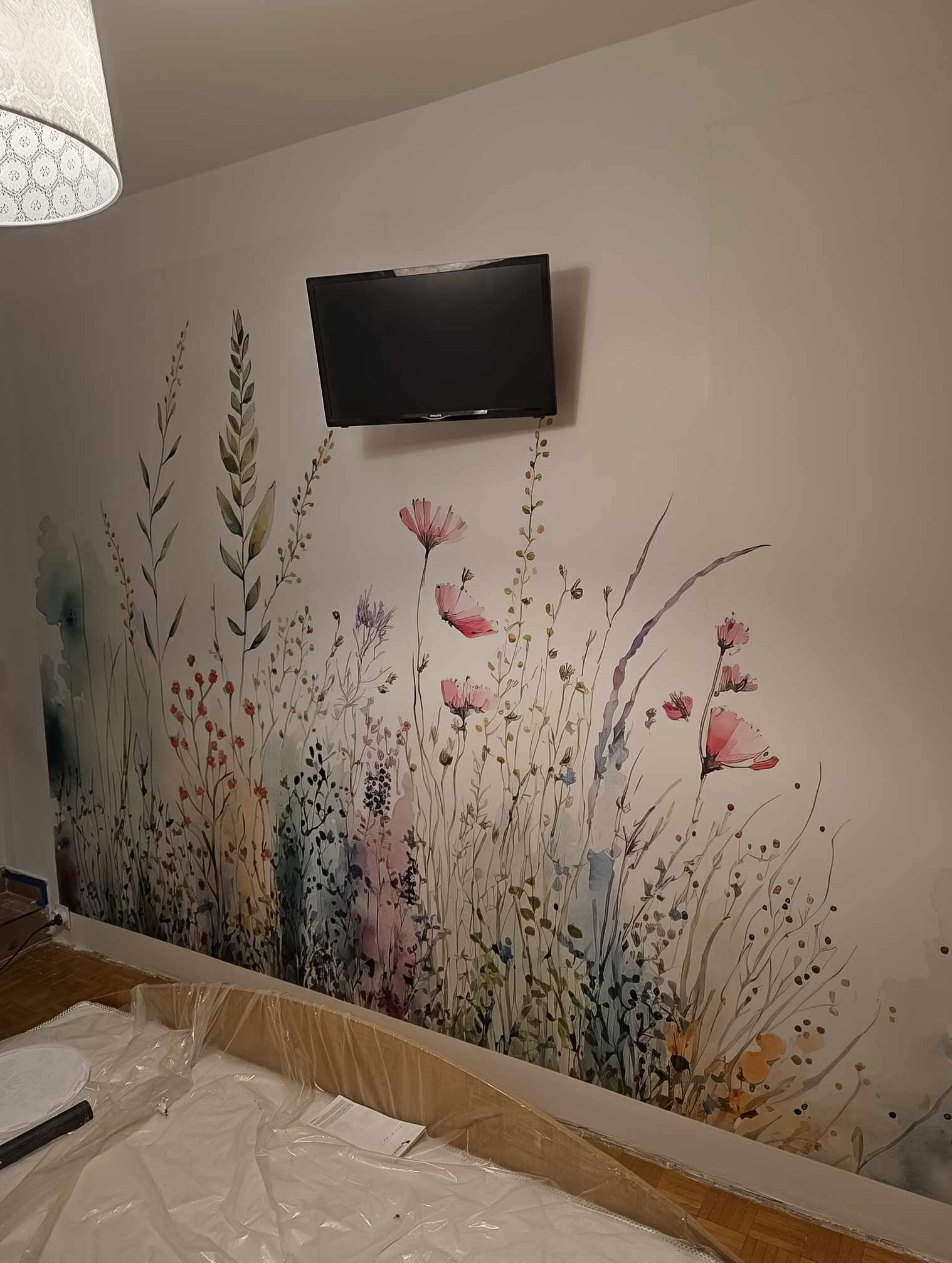 Ściana w pokoju zdobiona malowidłem ściennym przedstawiającym łąkę z kwiatami w stylu akwareli, z zawieszonym telewizorem na górze. Meble przykryte folią ochronną.