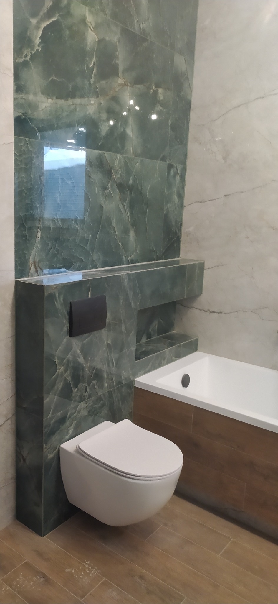 Nowoczesna łazienka z podwieszaną toaletą, zielonymi płytkami imitującymi marmur i zabudowaną wanną z drewnopodobnymi panelami.