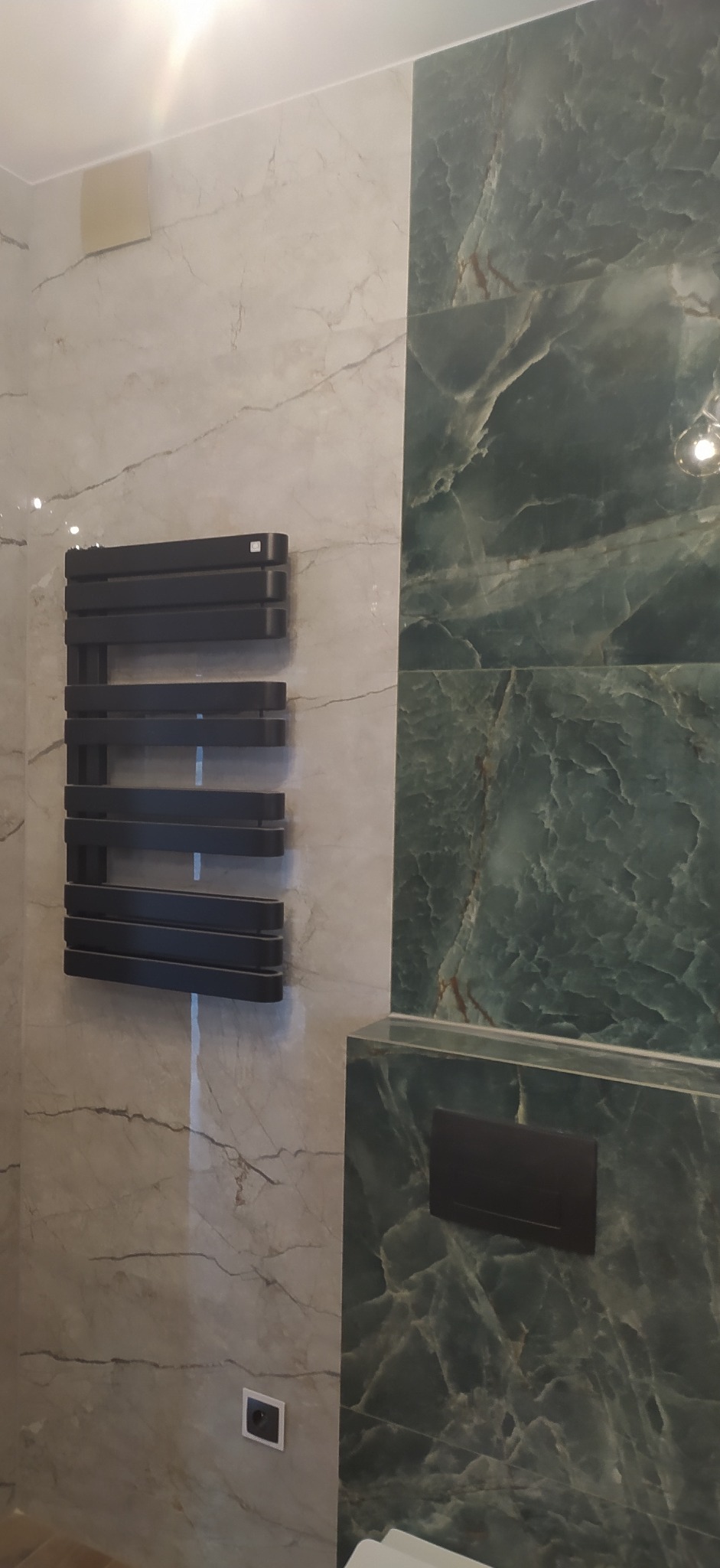 Nowoczesny, czarny grzejnik drabinkowy na ścianie z marmurowymi płytkami w odcieniach szarości i zieleni, widoczny panel spłuczki WC i gniazdko elektryczne.