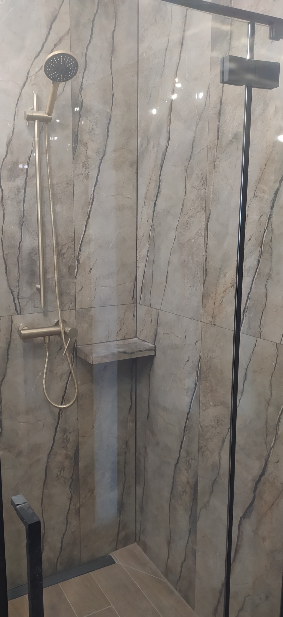 Kabina prysznicowa z deszczownicą w kolorze szczotkowanego złota, półka wbudowana w ścianę z płytek imitujących marmur.