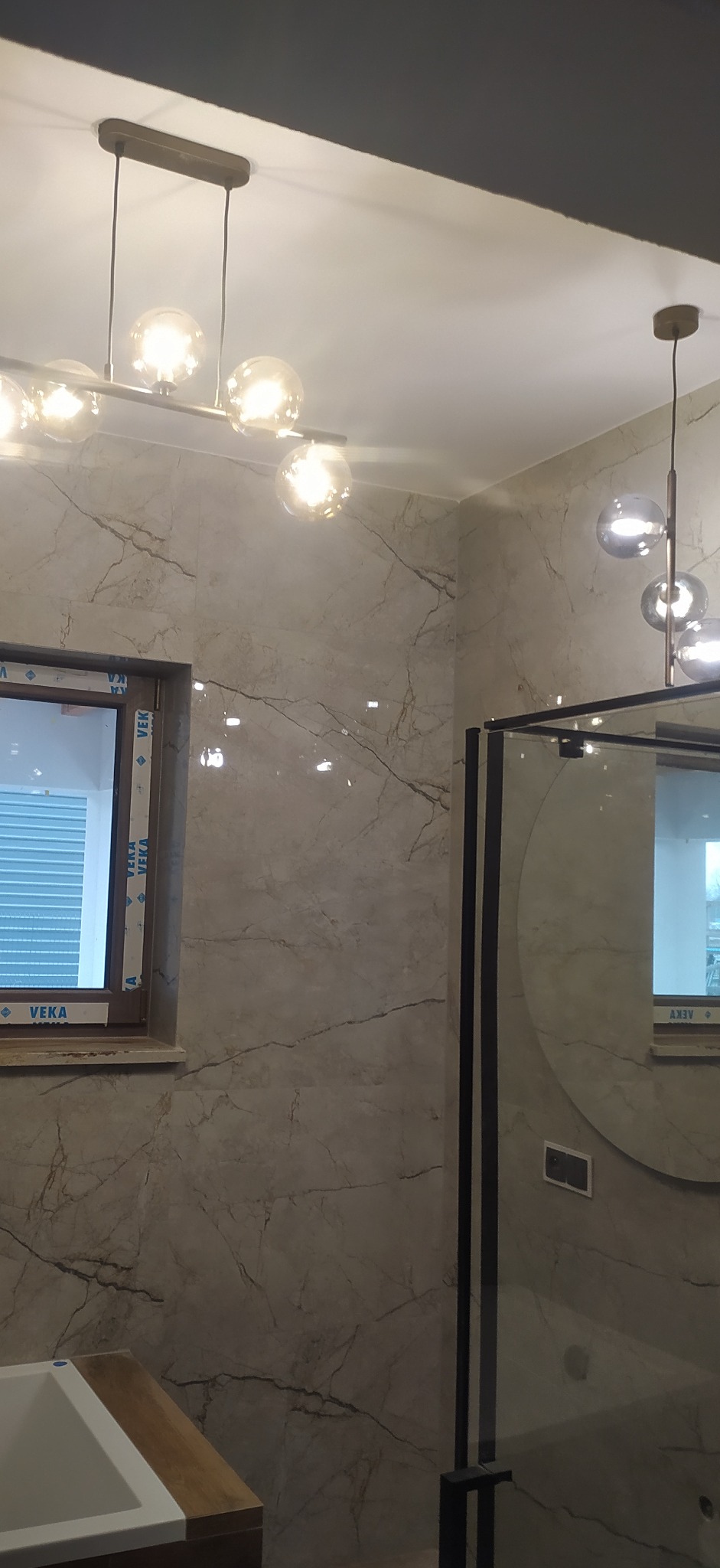 Wykończona łazienka z marmurowymi płytkami, nowoczesnym oświetleniem kulistym i czarną kabiną prysznicową z okrągłym lustrem.