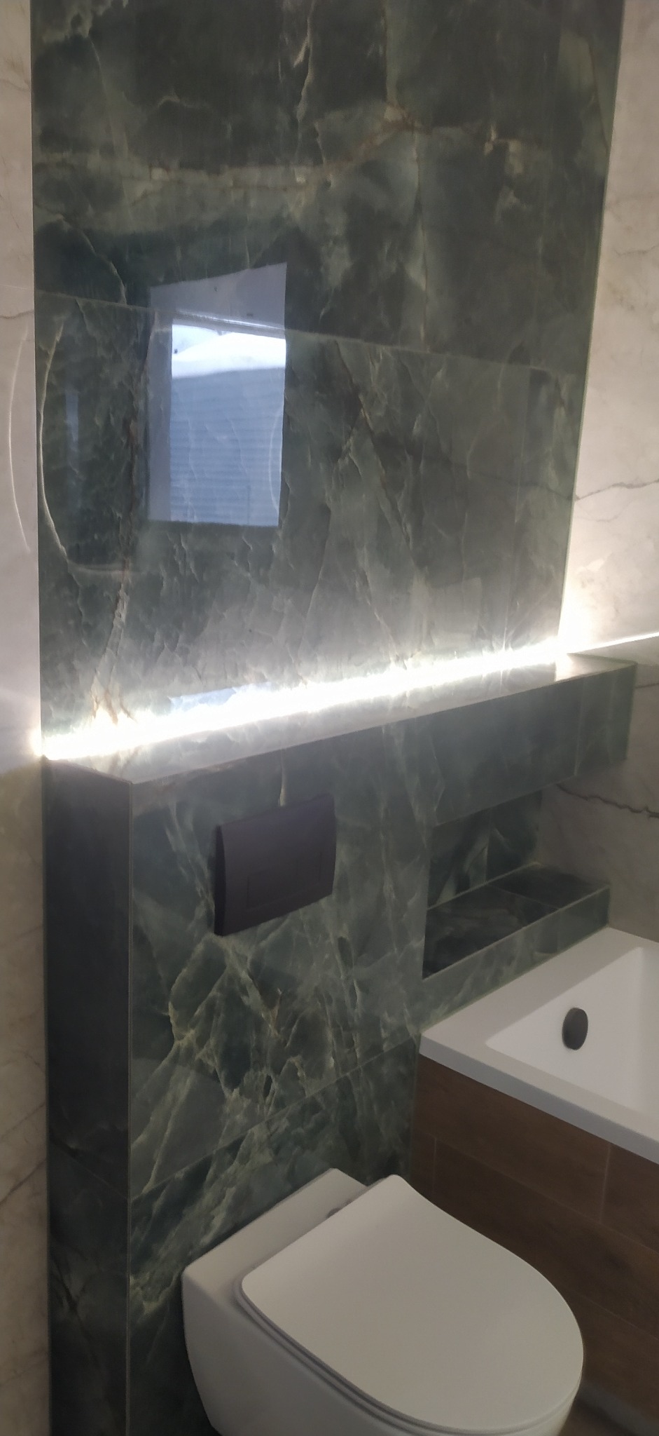 Ściana łazienki wyłożona zielonymi płytkami imitującymi marmur, podświetlona listwą LED, z widoczną toaletą i fragmentem wanny