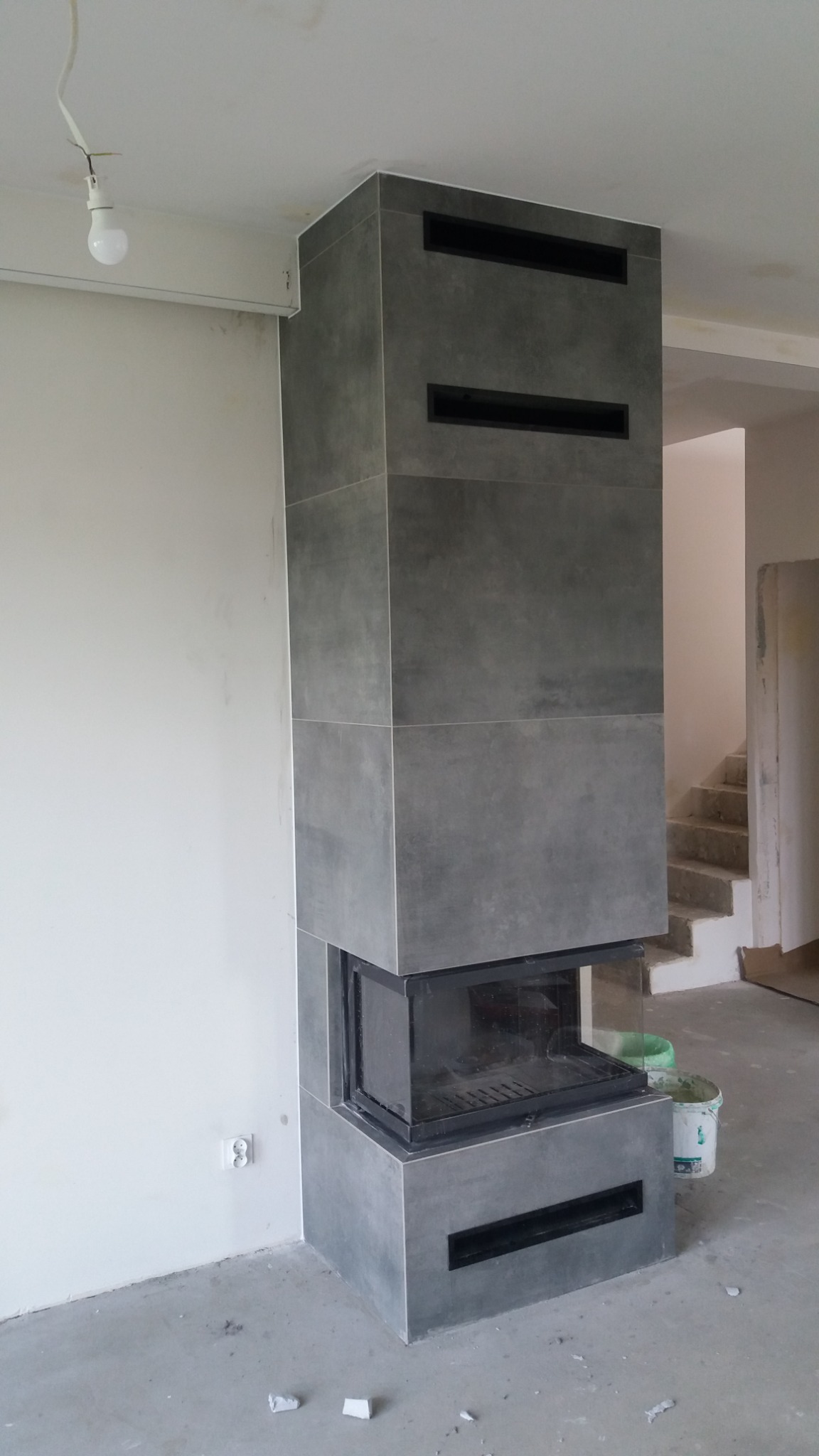 Obudowa kominka z szarych, kwadratowych płyt w surowym wnętrzu, widoczna puszka elektryczna na ścianie i schody w tle.