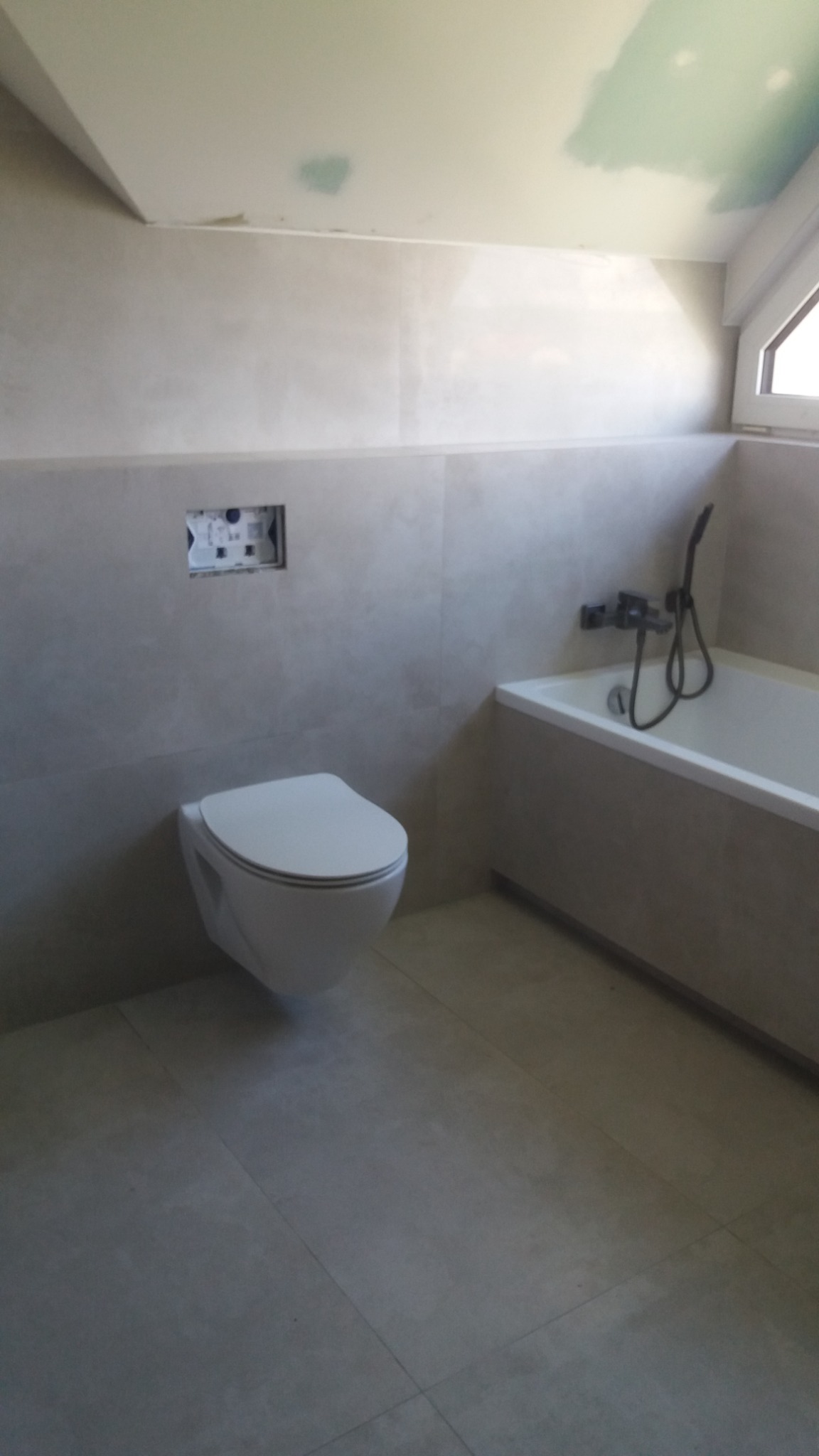 Nowoczesna łazienka w trakcie wykańczania, z podwieszaną toaletą, wanną i szarą glazurą, widoczne ślady prac na suficie.