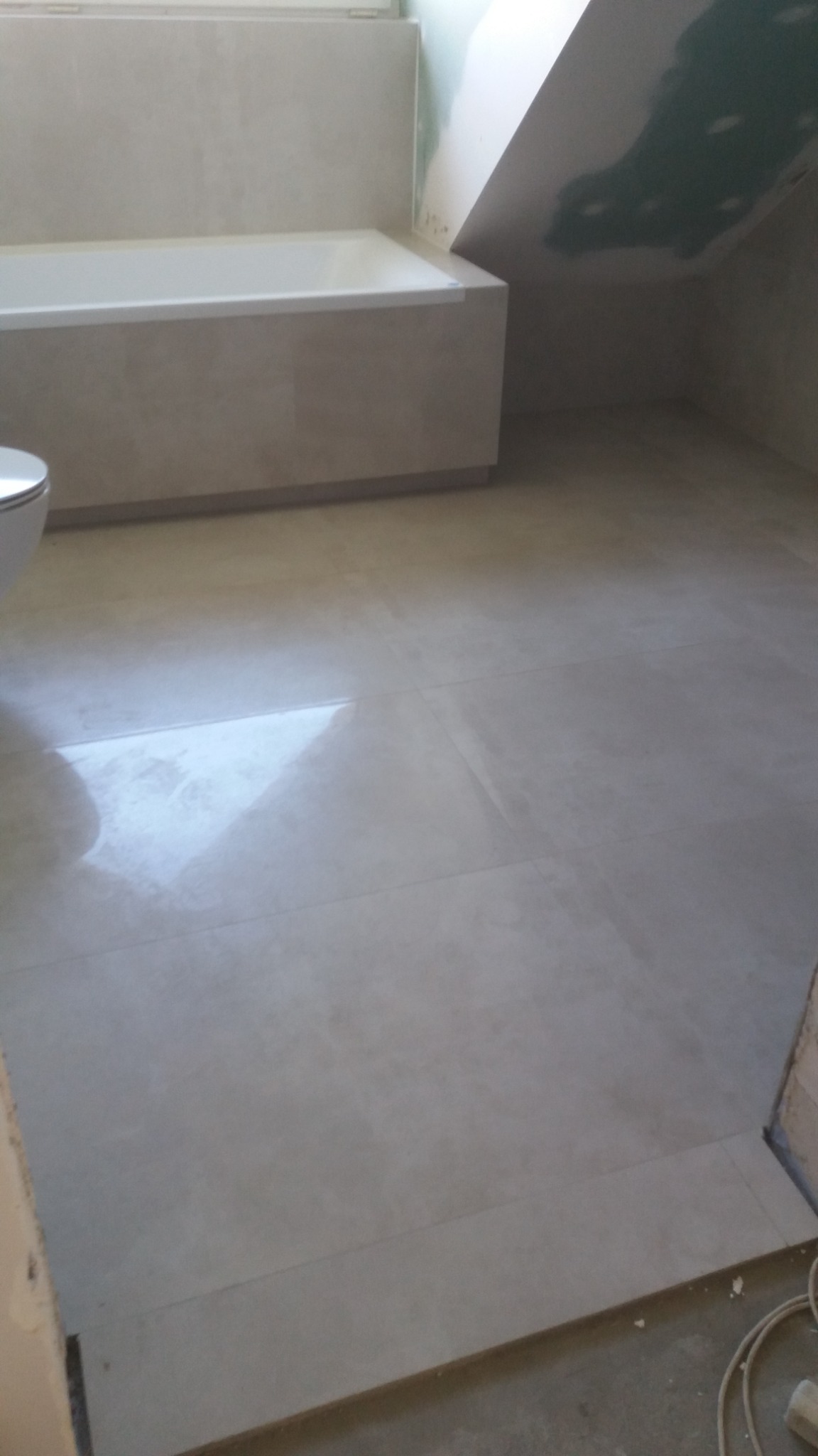 Jasne, błyszczące płytki podłogowe w łazience z wanną obudowaną płytkami i skośnym sufitem w tle, widoczne niedokończone prace na suficie.