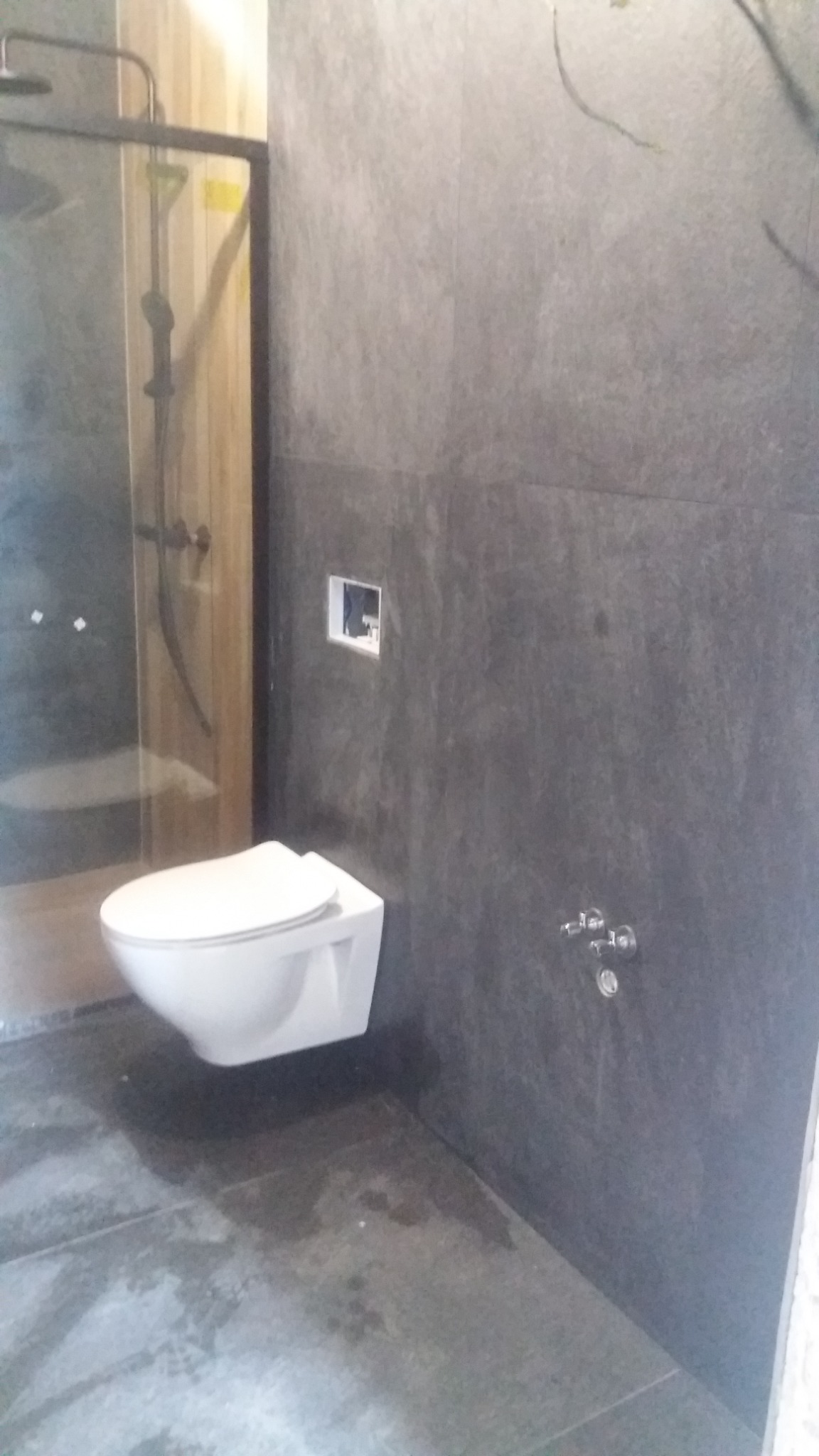 Nowoczesna łazienka z szarą okładziną ścienną i podwieszaną toaletą, kabina prysznicowa w tle, widoczny fragment instalacji wodnej.