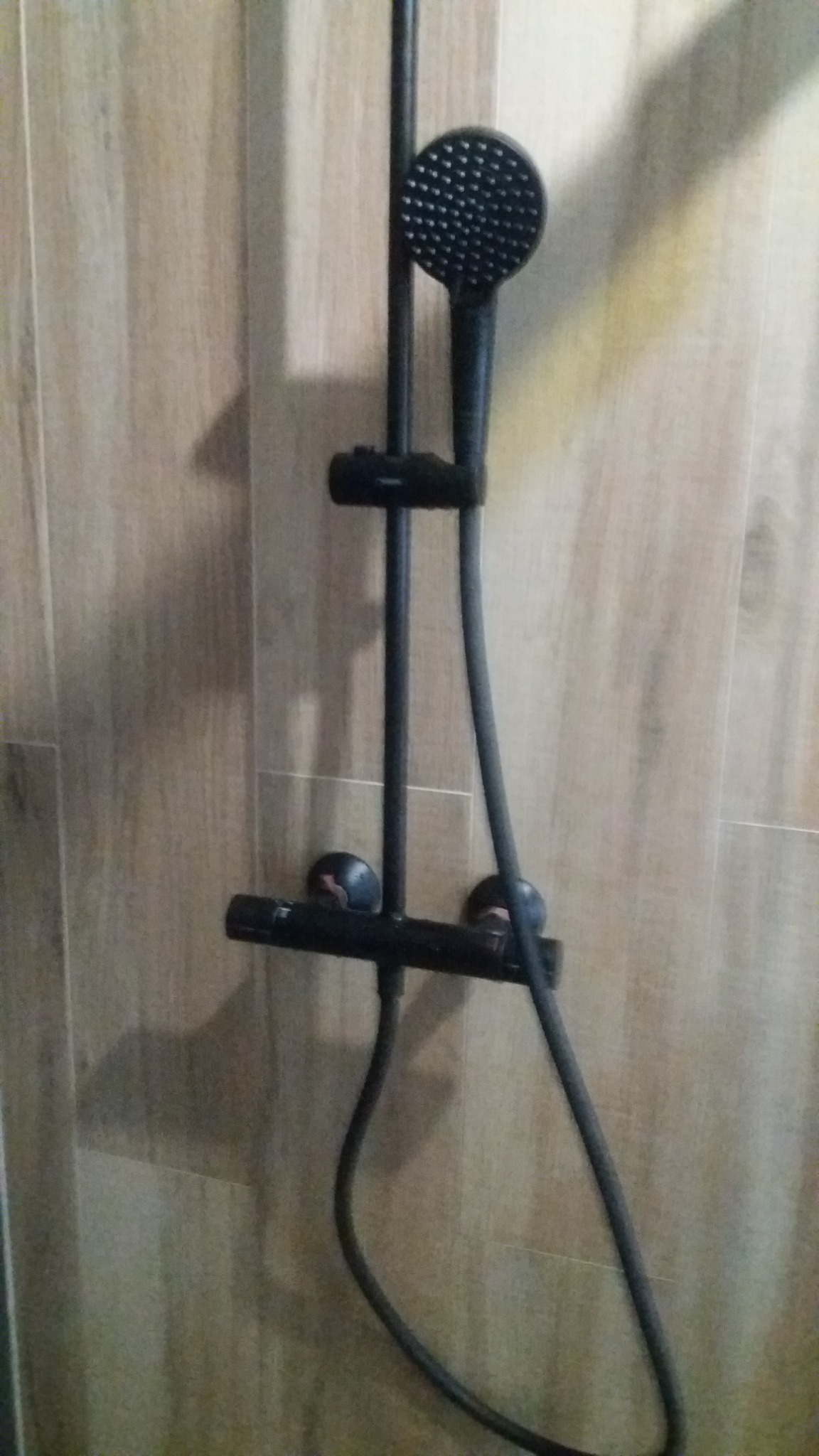 Czarny panel prysznicowy z deszczownicą i termostatem, zamontowany na ścianie wyłożonej panelami imitującymi drewno.