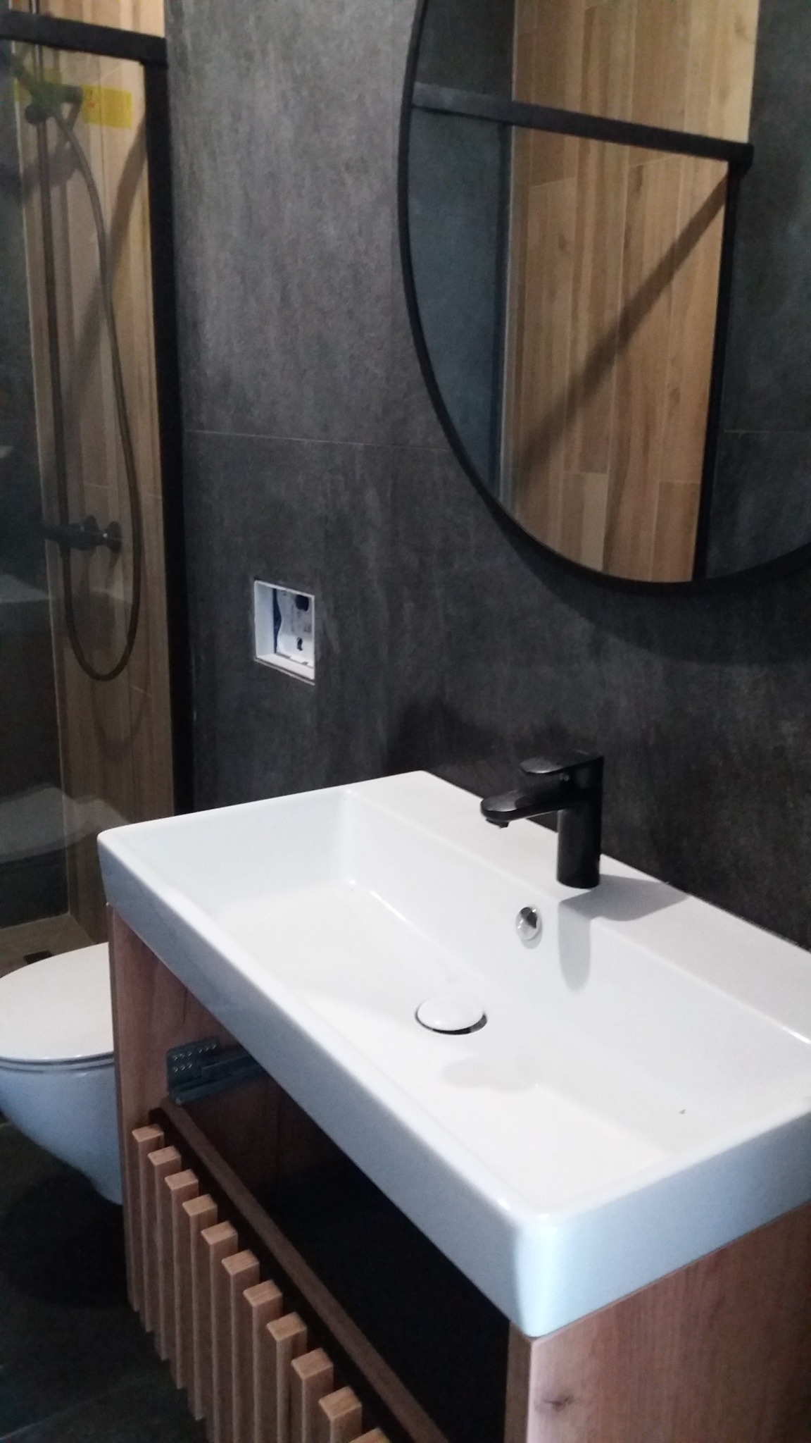 Nowoczesna łazienka z umywalką nablatową, czarną armaturą i okrągłym lustrem na tle szarych płytek oraz kabiny prysznicowej.