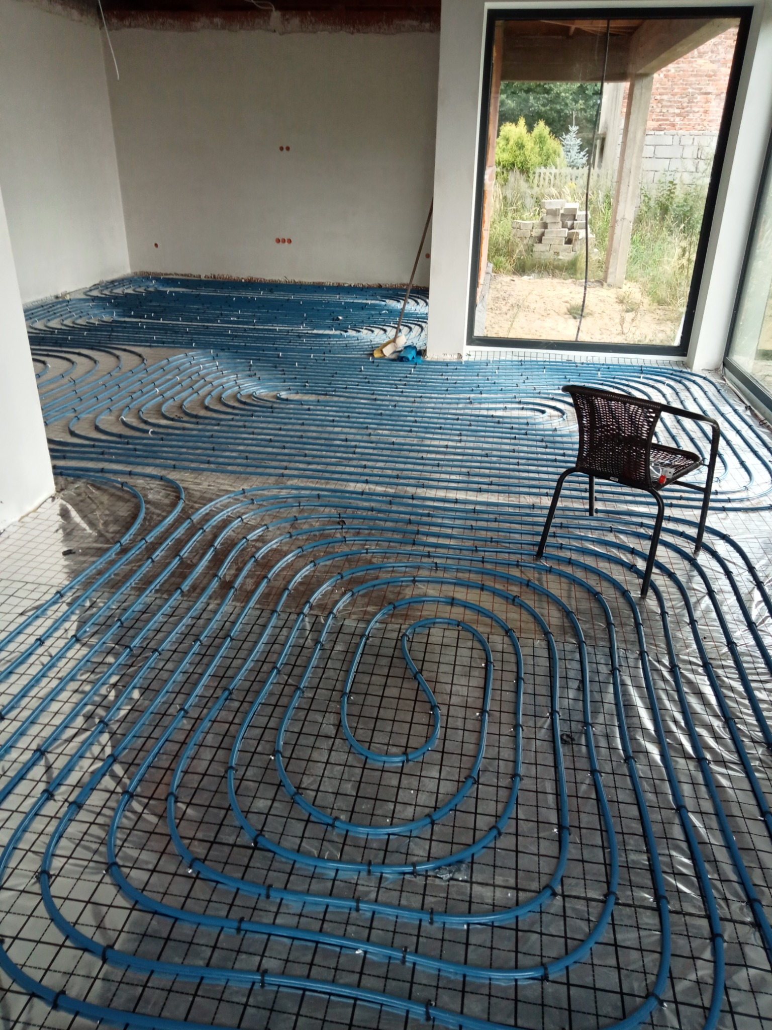 Niebieskie rury ogrzewania podłogowego ułożone spiralnie na folii w surowym wnętrzu, z krzesłem na pierwszym planie i widokiem na ogród przez duże okno.