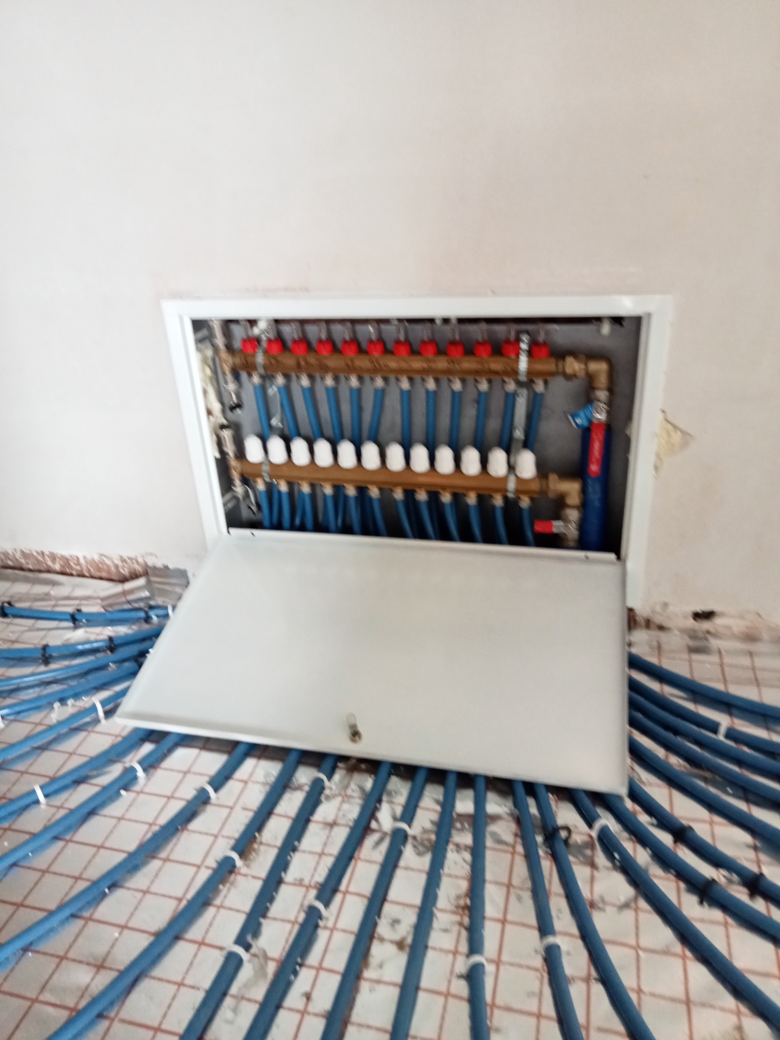 Rozdzielacz ogrzewania podłogowego w szafce, z widocznymi niebieskimi rurami ułożonymi na podłodze z siatką, przed wylaniem posadzki.