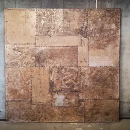 Dekoracyjny panel ścienny w stylu industrialnym, złożony z metalowych płyt o zróżnicowanej fakturze i odcieniach brązu, połączonych nitami na tle surowego, betonowego muru.