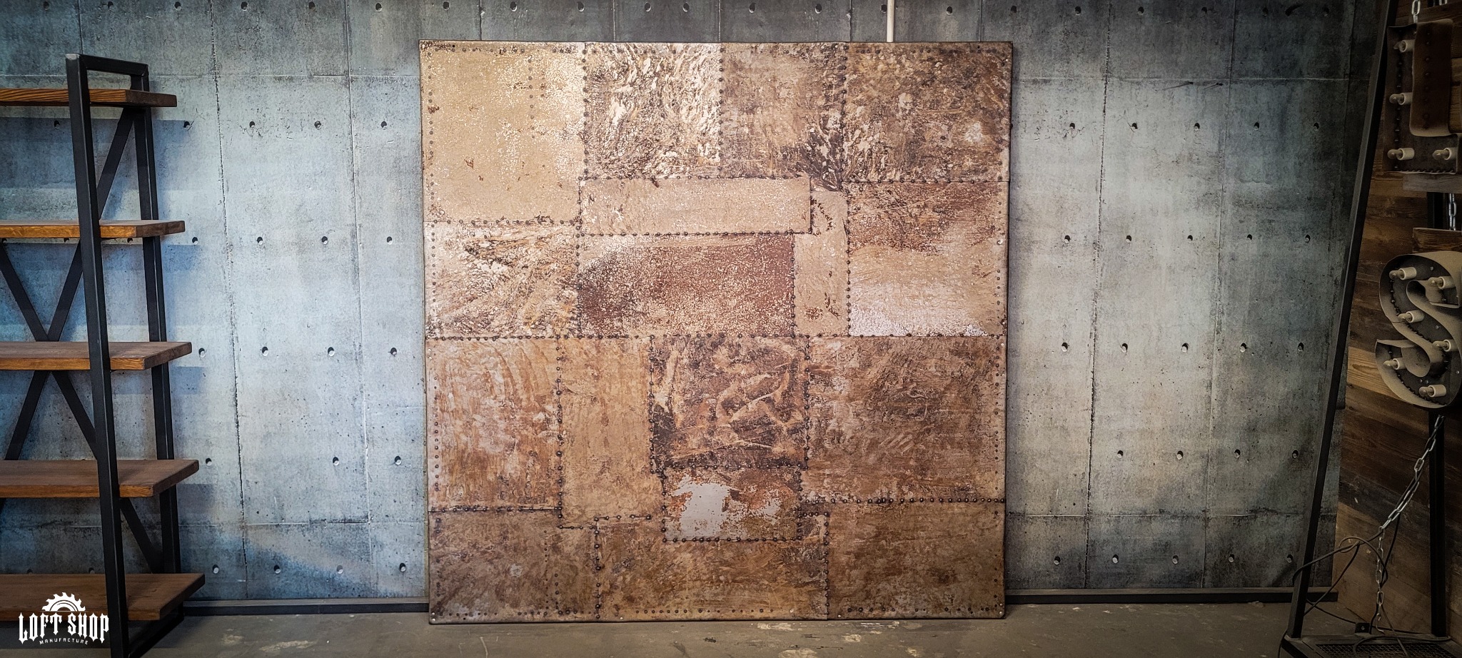 Dekoracyjny panel ścienny w stylu industrialnym, złożony z metalowych płyt o zróżnicowanej fakturze i odcieniach brązu, połączonych nitami na tle surowego, betonowego muru.