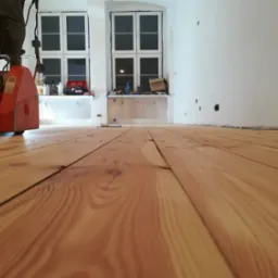 Szlifowanie drewnianej podłogi parkieciarką marki Scorpion w jasnym pomieszczeniu z oknami, widoczne ślady po szpachlowaniu przy ścianach.