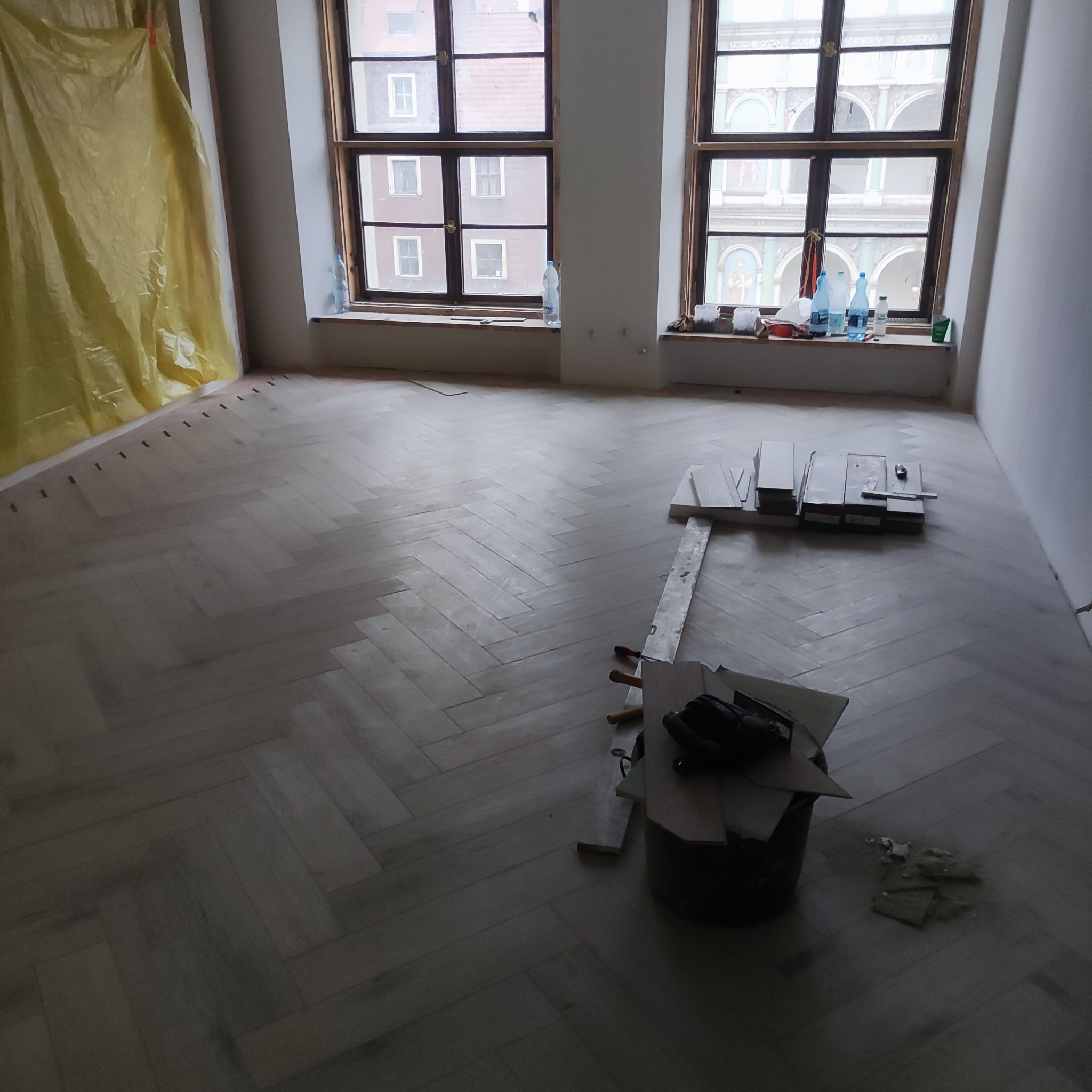 Układanie jasnej, drewnianej podłogi w jodełkę francuską w przestronnym pokoju z dwoma oknami. Część podłogi zabezpieczona folią ochronną, widoczne narzędzia i resztki materiałów w wiadrze.