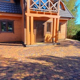 BrukPol Wiesław Kraiński - Drewniany dom z brązowymi oknami i balkonem, z wyłożonym kolorową kostką brukową podjazdem.