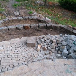 BrukPol Wiesław Kraiński - Układanie kamiennej ścieżki w ogrodzie, widoczne obrzeża z kostki granitowej, stos kamieni do dalszej pracy i zielona konewka.