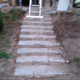 BrukPol Wiesław Kraiński - Kamienne schody zewnętrzne w trakcie budowy, widoczna drabina oparta o stopnie, ziemia i roślinność w tle.